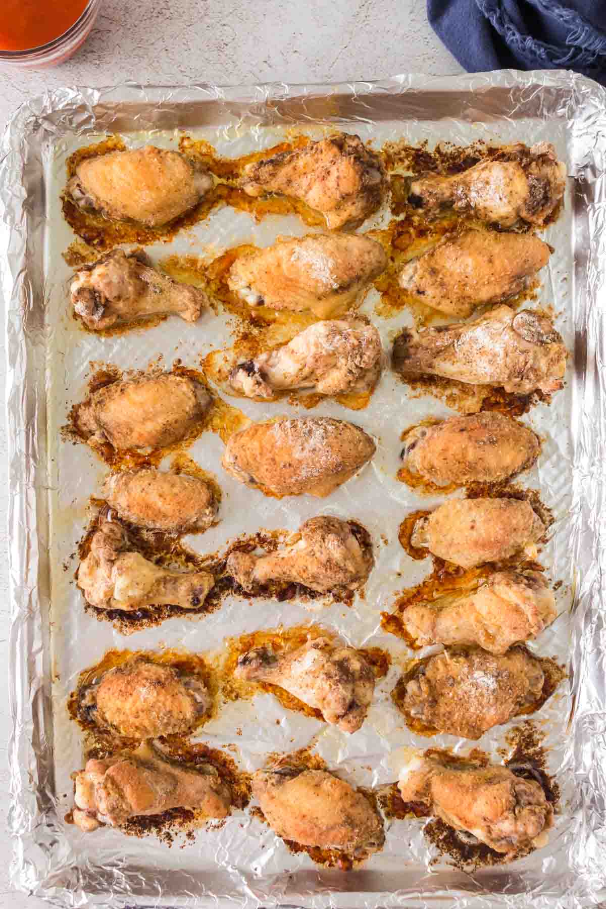 A foil-lined baking tray filled with cooked chicken drumettes, arranged in rows. The chicken is seasoned and roasted, with a crispy texture visible on the surface. The tray is resting on a light-colored surface.