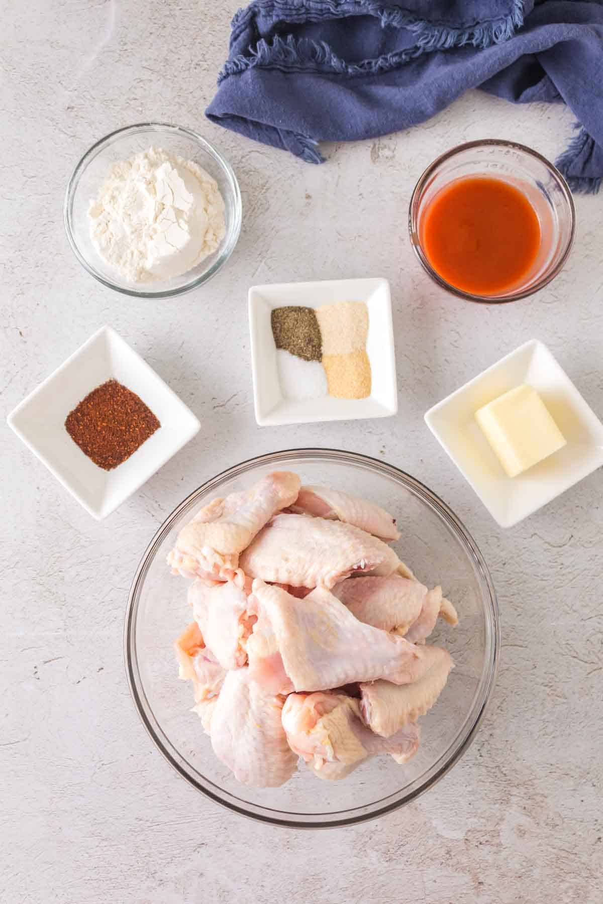 A bowl of raw chicken wings is surrounded by small dishes containing flour, hot sauce, butter, and seasonings including salt, pepper, garlic powder, and paprika. A blue cloth is placed at the top of the image on a light-colored surface.