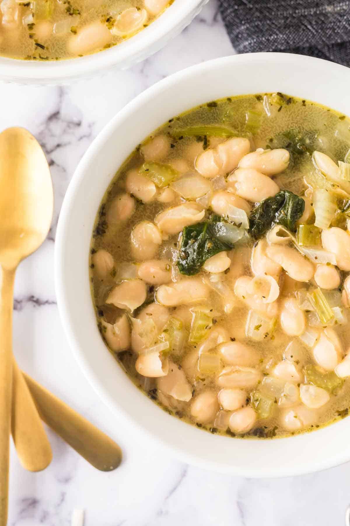 A bowl of white bean and spinach soup on a marble surface. The soup includes white beans, spinach, and chopped herbs, with a golden spoon placed beside the bowl. The broth appears creamy and seasoned. Another partial bowl is visible at the top.