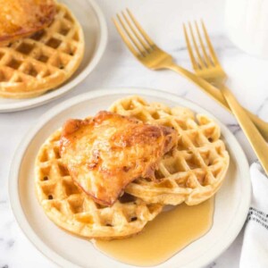 Plates of chicken and waffles, made from a beloved chicken and waffles recipe, are topped with syrup on a marble countertop. Two forks rest beside the dish, and a white and black striped napkin is nearby. Another plate with the same delicious meal is in the background.