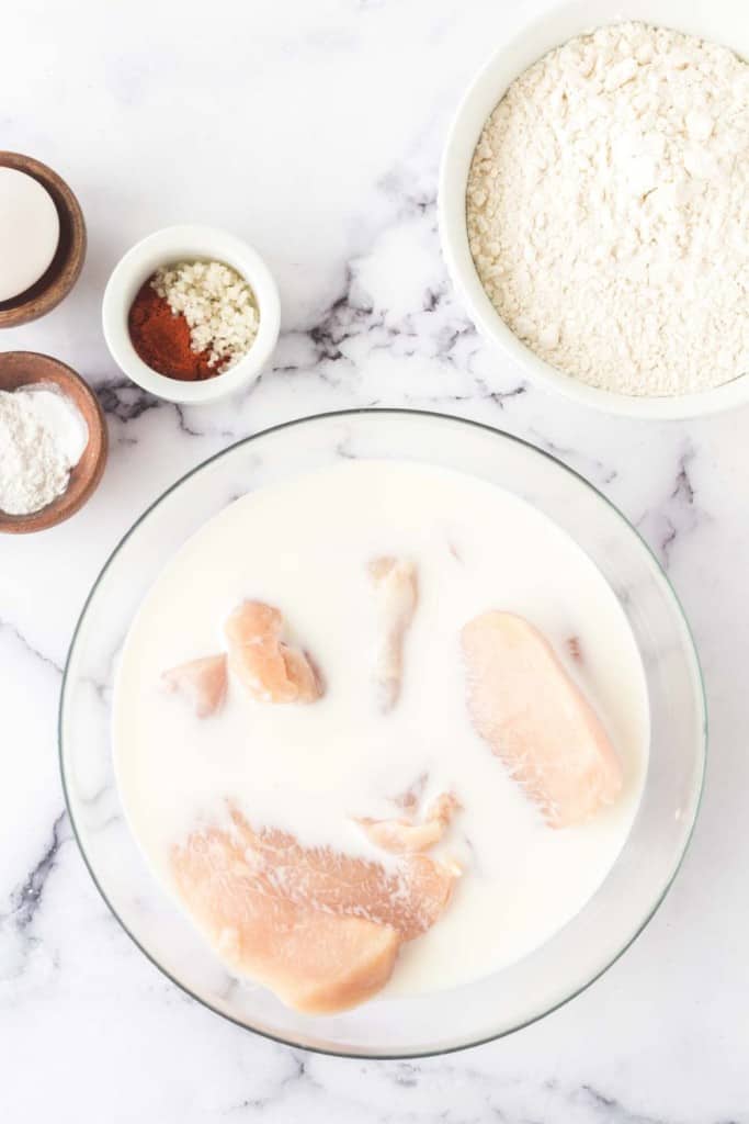 Raw chicken pieces soaking in a bowl of milk on a marble surface. Surrounding the bowl are ingredients in small bowls: flour, minced garlic, paprika, and a single egg.