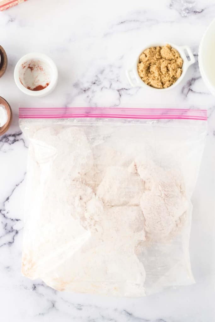 A plastic ziplock bag filled with a flour mixture on a marble surface. Nearby are small bowls containing brown sugar, a white powder, and a red spice.