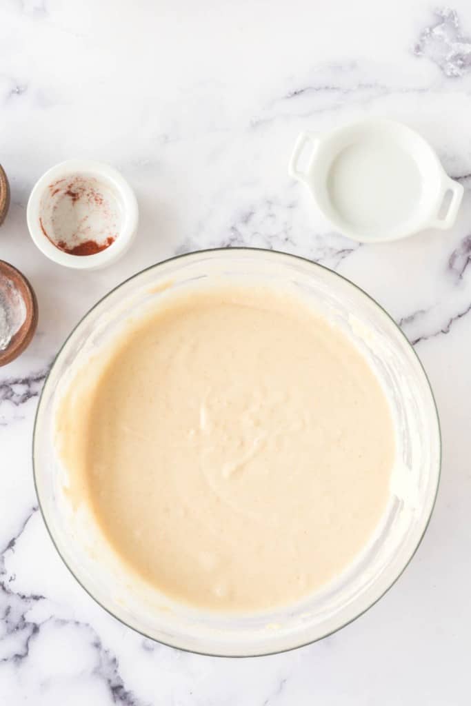 A glass bowl filled with creamy cake batter sits on a marble countertop. Surrounding it are small bowls containing red powder, flour, and a white liquid. The ingredients are ready for mixing or baking.