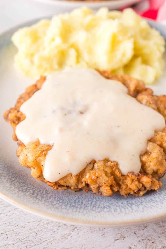 A plate featuring chicken fried steak smothered in creamy white gravy sits beside a generous serving of mashed potatoes. This classic dish is elegantly presented on a light-colored ceramic plate atop a pristine white surface.