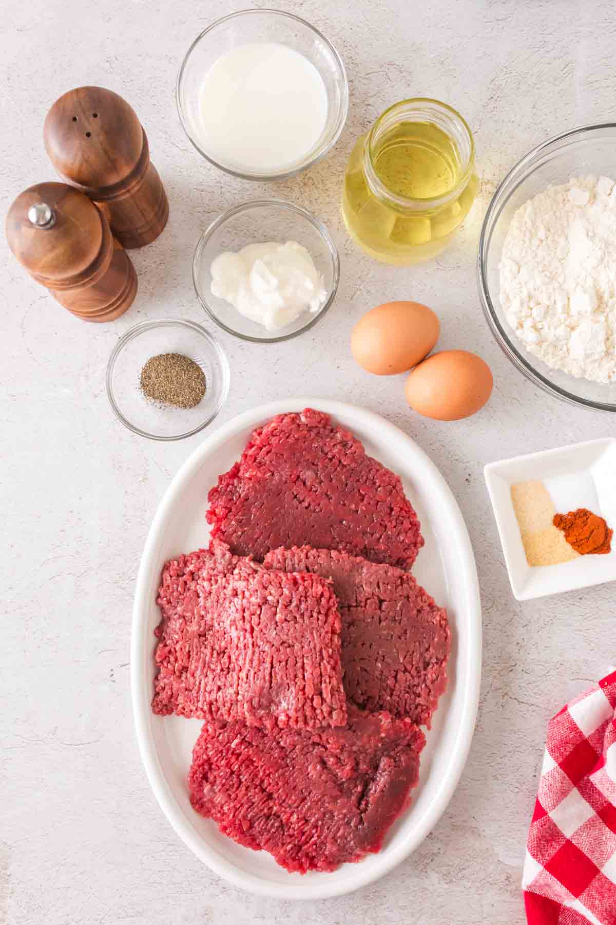 A white oval plate with raw cube steaks, perfect for making Chicken Fried Steak, sits on the countertop. Surrounding it are flour, eggs, milk, oil, sour cream, and seasonings like salt, pepper, paprika, and garlic powder. A red checkered cloth adds a charming touch at the bottom right.