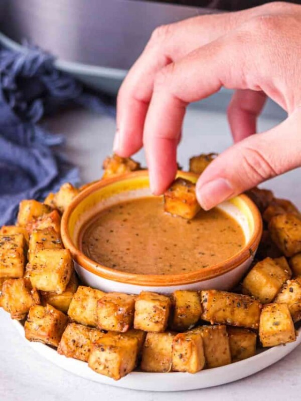 POV hand dipping into a plate of air fryer tofu with a dish of sauce in the center