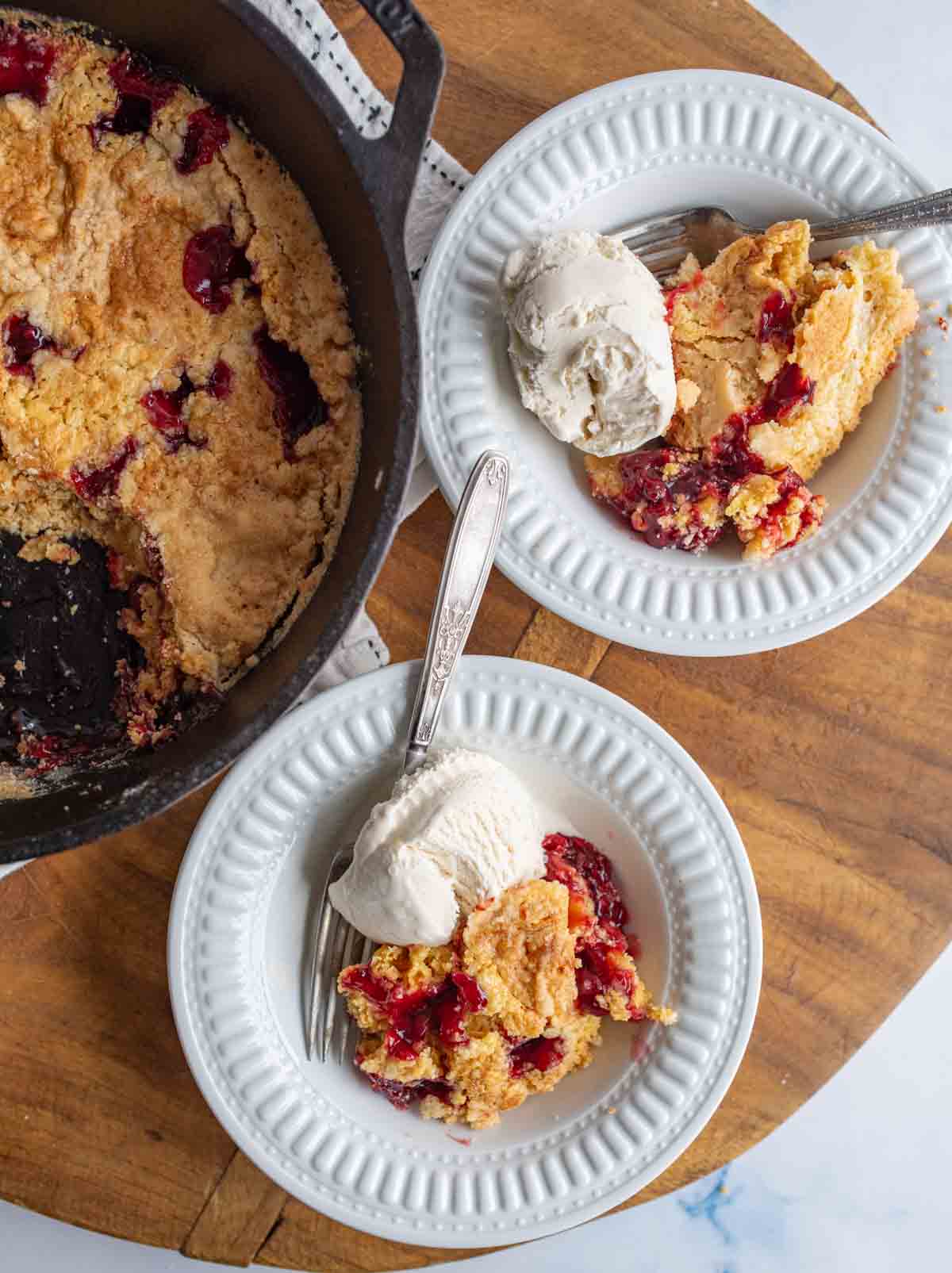 cherry dump cake in a cast iron pan with red filling peaking through a crumbly baked crust and two plate servings.