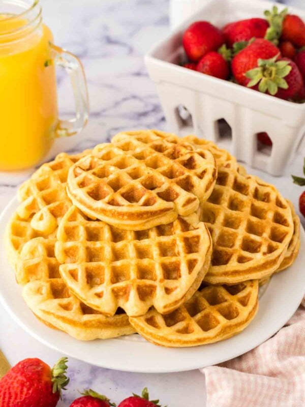 Waffles from a buttermilk waffle recipe, stacked on a white platter, surrounded by fresh strawberries. A glass mug of orange juice and a basket of strawberries grace the background, all set on a sleek marble surface.