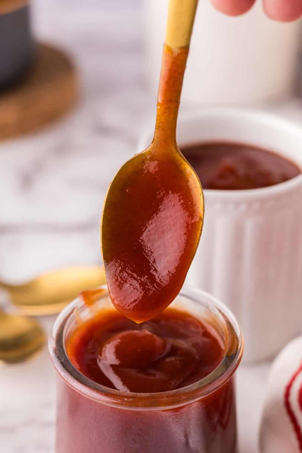small glass jar with barbecue sauce and a golden spoon being pulled out of it dripping sauce off of it.