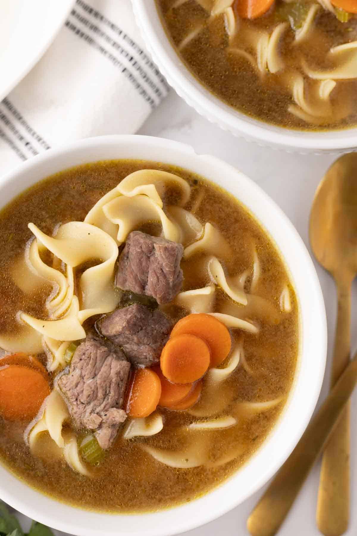 beef noodle soup recipe served in white soup bowls with a wooden spoon to the side.