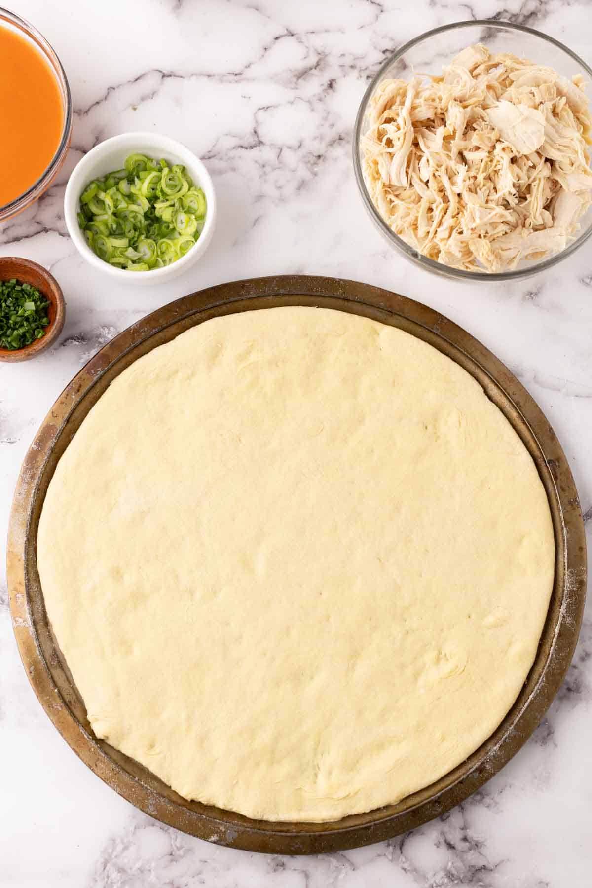 round cooking stone with raw pizza dough surrounded by ingredients to make buffalo pizza.