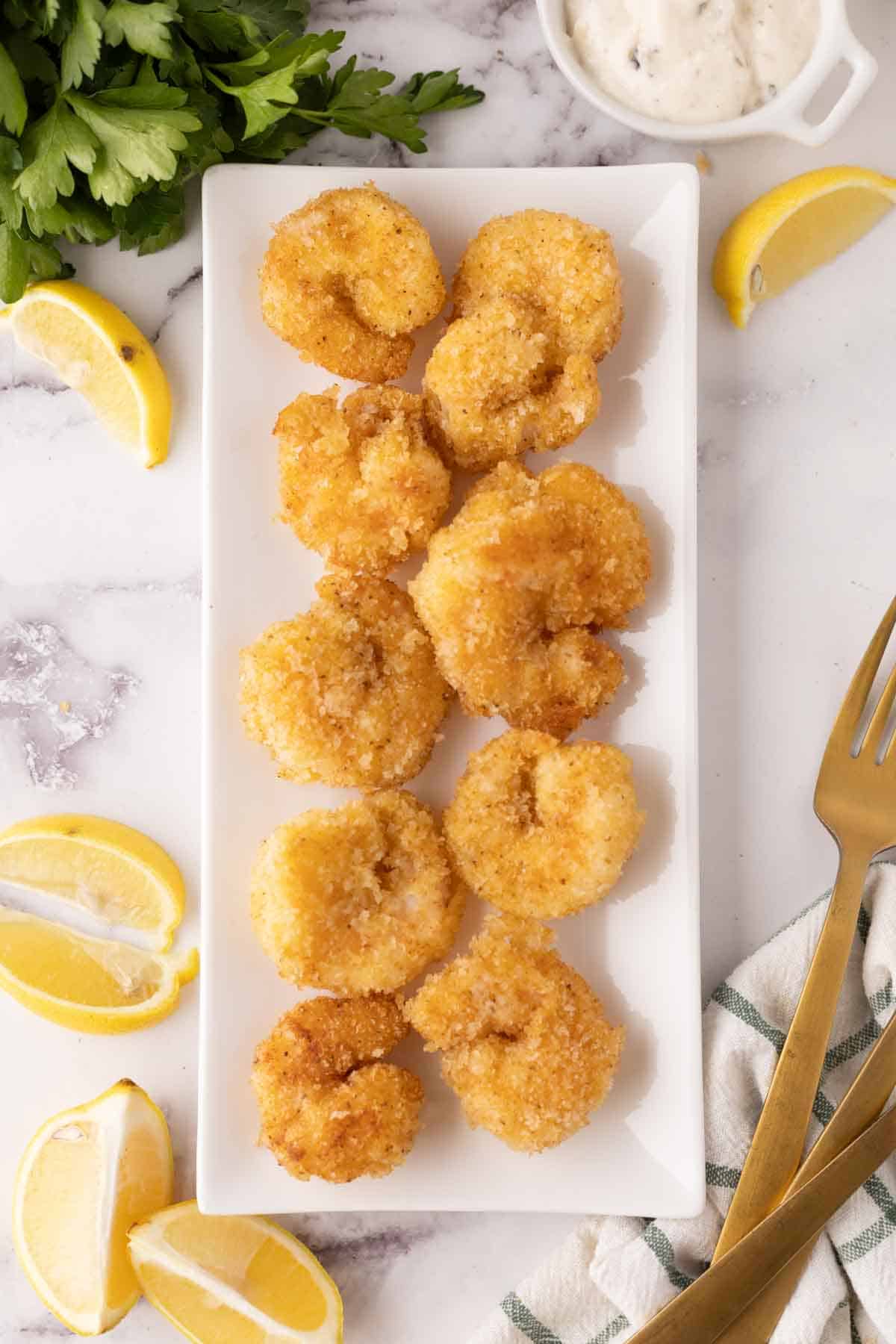 two rows of fried butterfly shrimp on a white rectangle plate with lemons to the side.