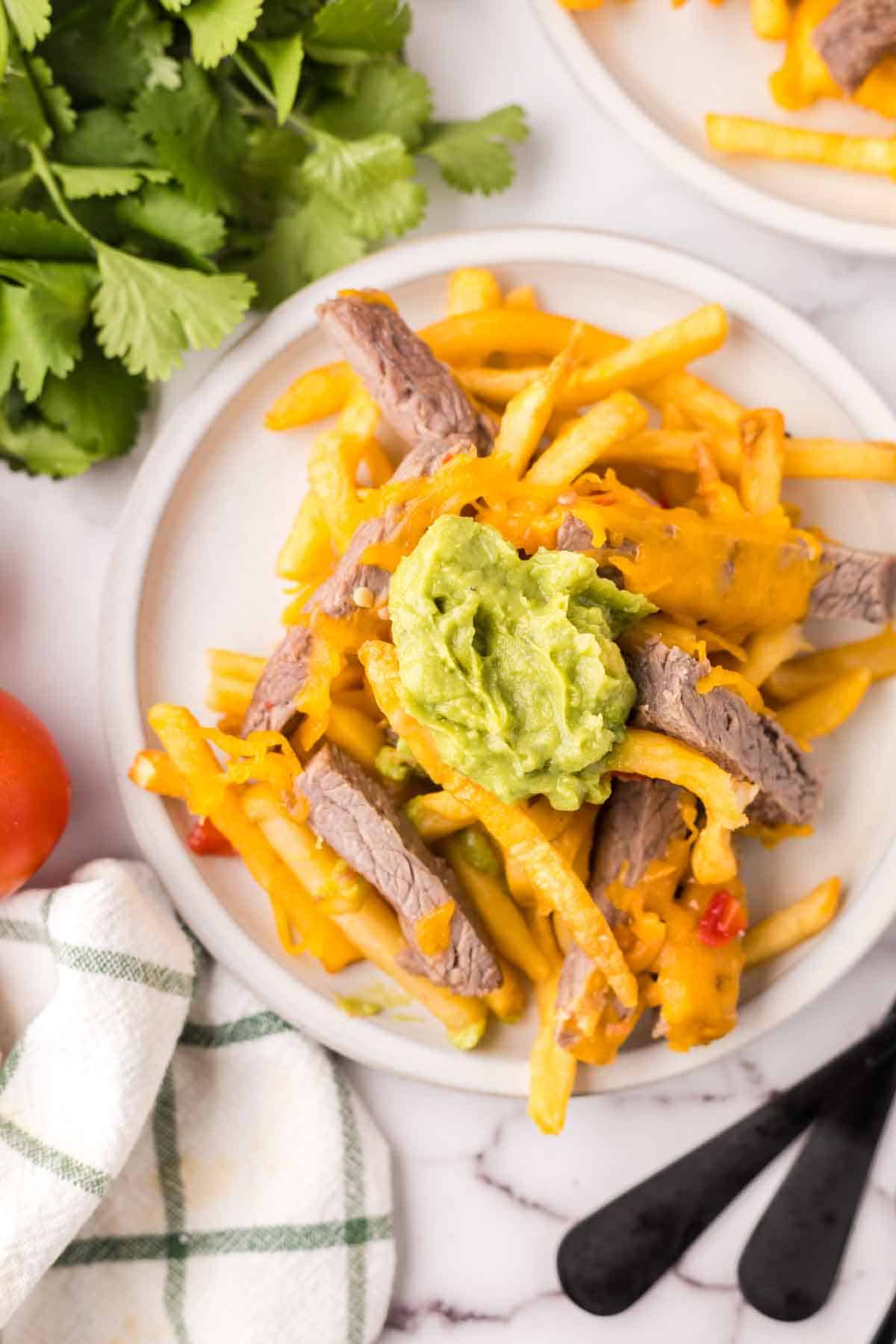 carne asada fries on a round white plate with black forks and tomato to the side.