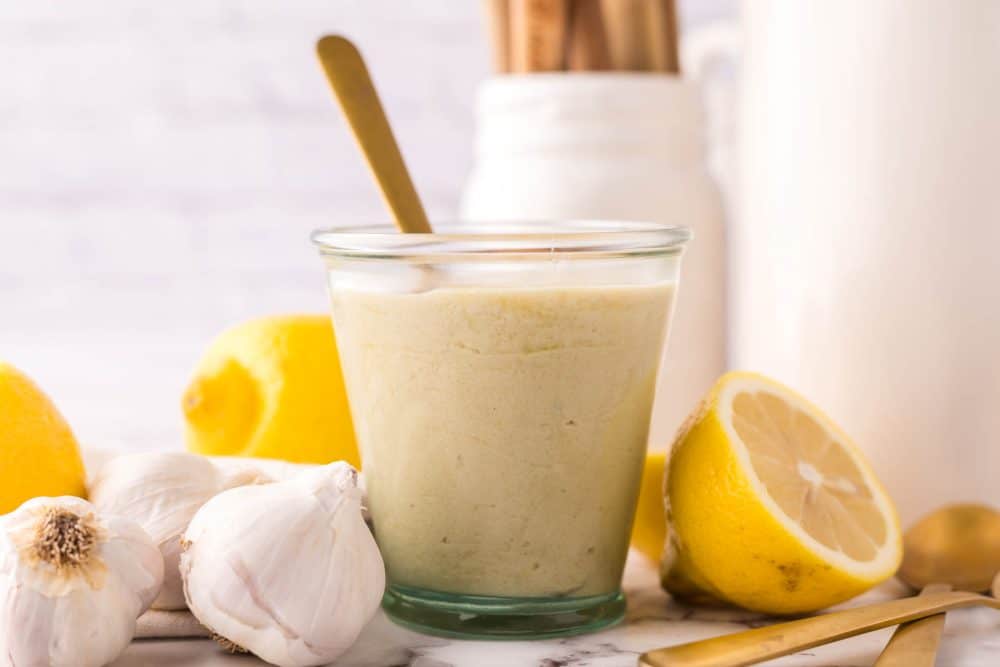 tahini dressing in a small glass jar with lemons and garlic all around with a gold spoon in it.