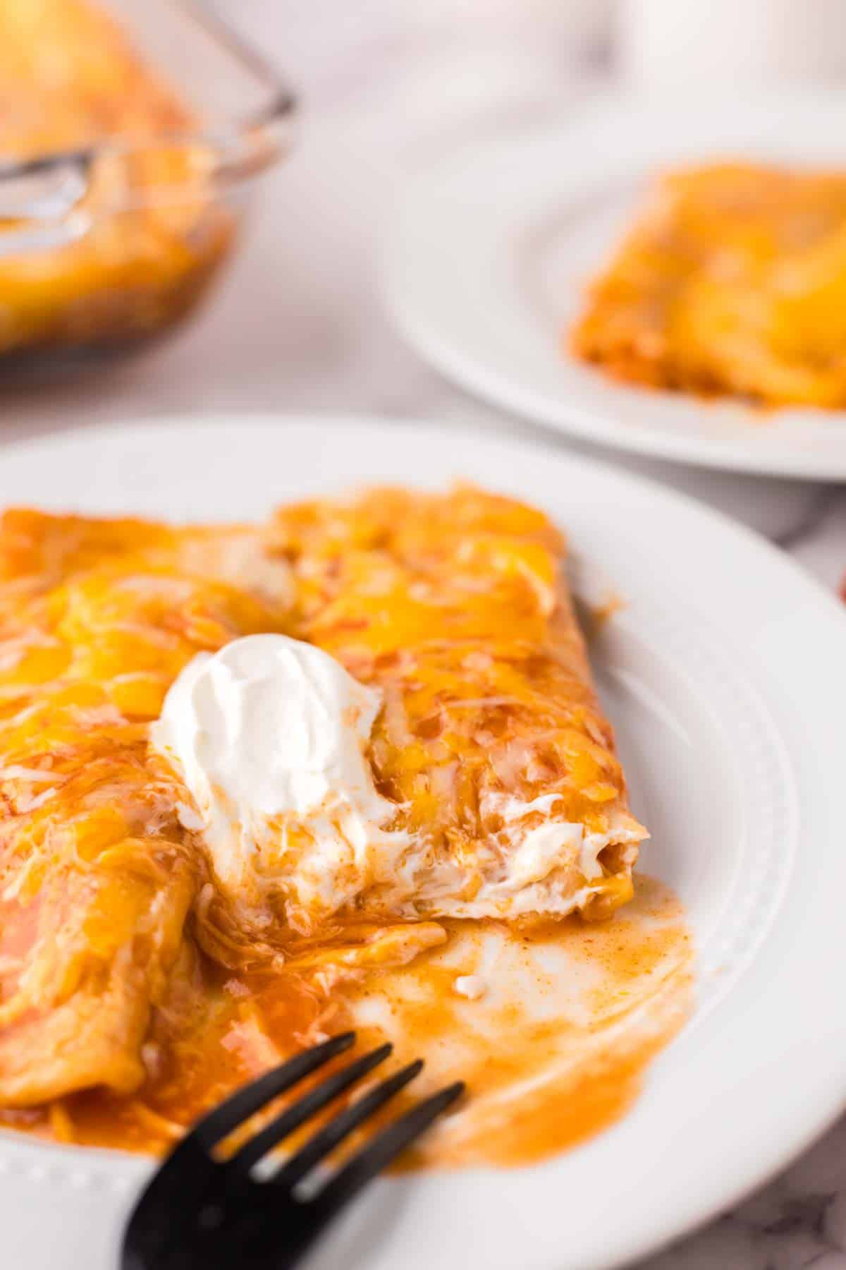 cheese enchiladas on a white plate with sour cream on top with a bite taken out.