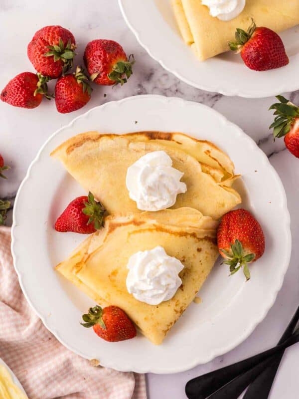 folded crepes on a white plate with fresh berries around and whipped cream.