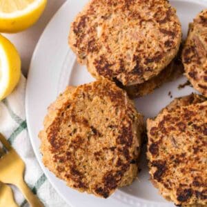 four tuna patties on a round white plate with lemons to the side.