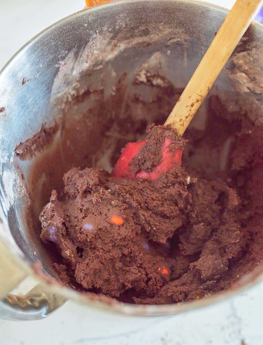 Dark chocolate Halloween M&M Cookie dough in a mixing bowl with a red spatula with a wooden handle mixing in the M&Ms.