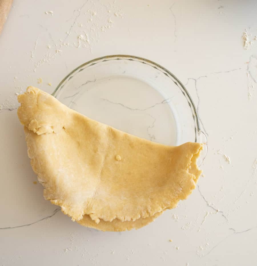 The pie crust unfolded into a semi-circle in a clear glass pie plate on a white counter. 