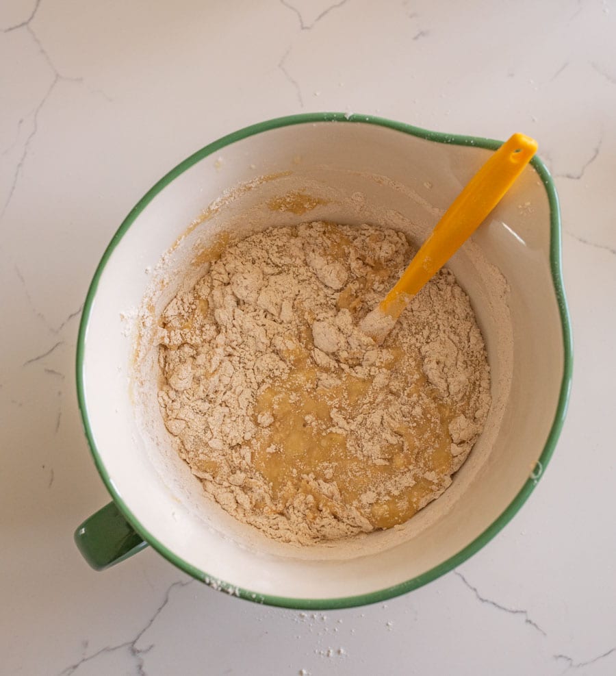 Sourdough banana bread batter with a yellow spatula used to stir in the dry ingredients in a white bowl with a green rim on a white counter. 