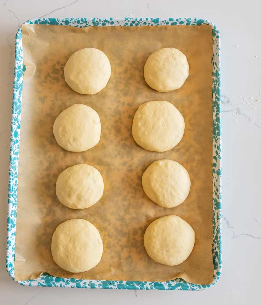 Eight sourdough hamburger buns on brown parchment paper on a blue and white speckled enamelware pan ready to go into the oven. 