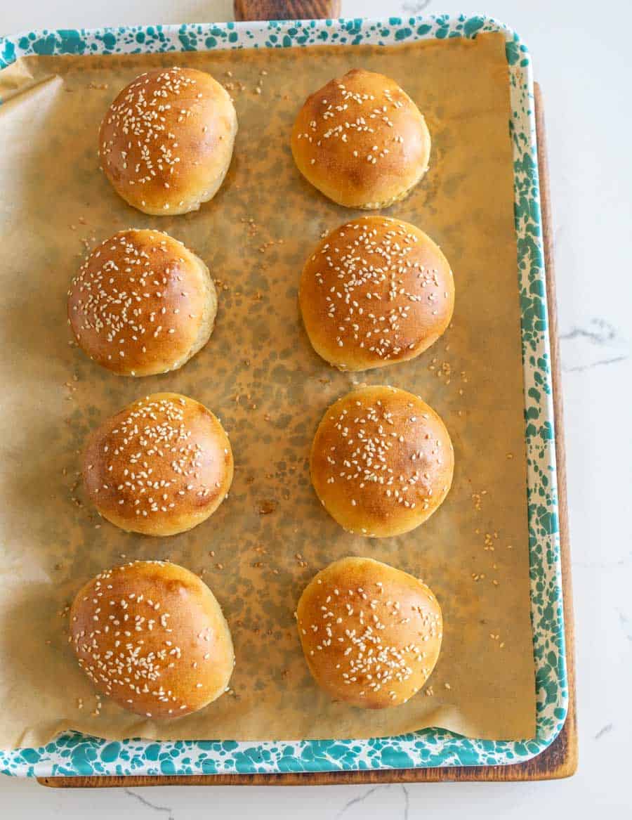 Eight freshly baked golden brown sourdough hamburger buns with sesame seeds on parchment paper on a blue and white enamelware pan. 