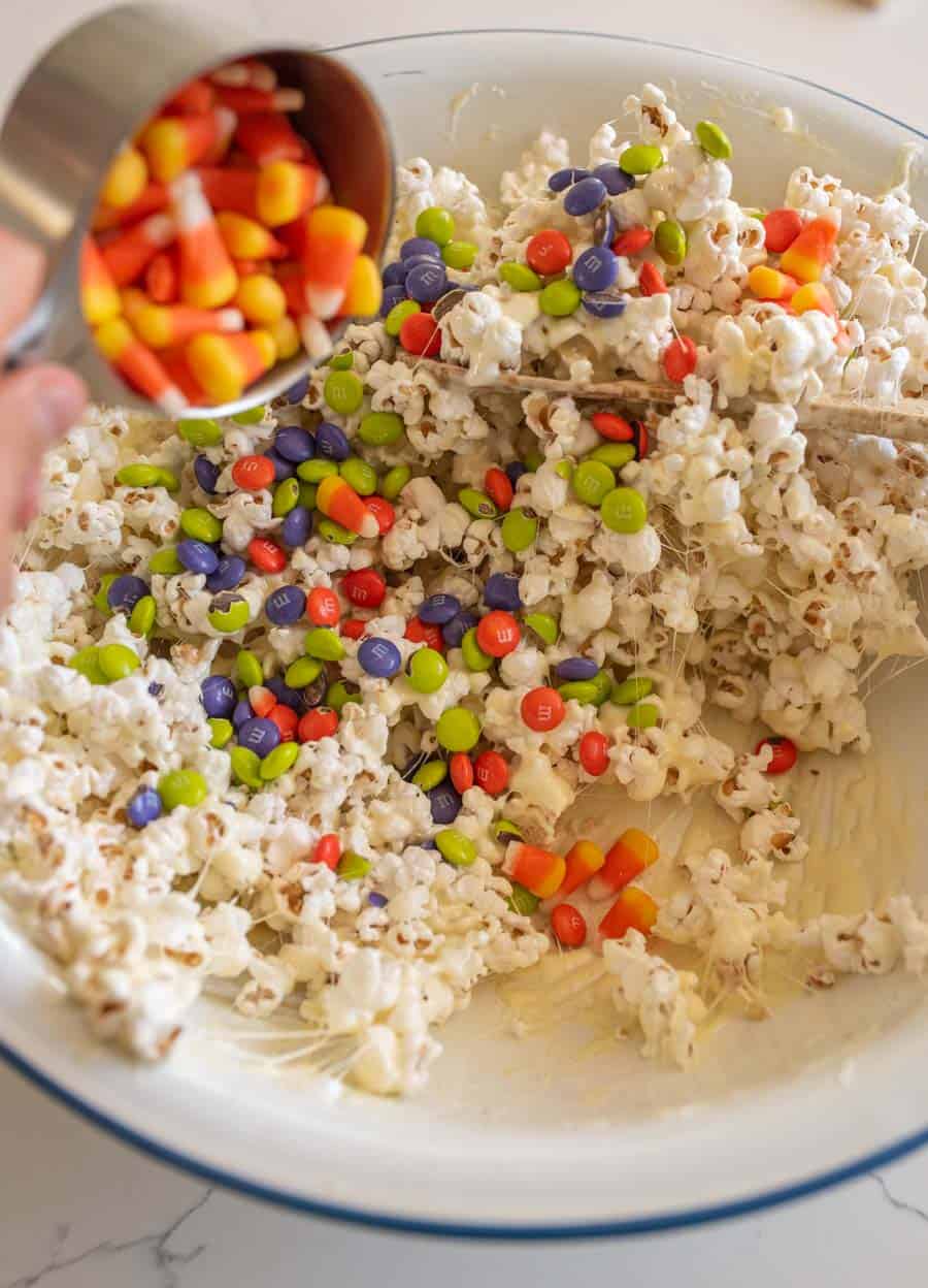 White bowl with popcorn coated in the melted marshmallow mixture with M&Ms and a small measuring cup pouring in the candy corn. 
