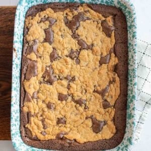 A speckled blue baking dish holds a cheater brookies recipe featuring a freshly baked chocolate and cookie crumble dessert. The golden brown cookie topping sits enticingly over the rich, dark chocolate base. This delightful treat rests on a wooden board with a white cloth nearby.