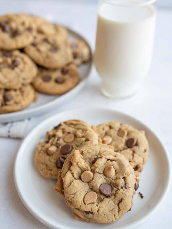 A small plate with three peanut butter chocolate chip cookies sits in the foreground, accompanied by a glass of milk. In the background, more cookies are piled on a larger plate. The scene is set on a light-colored surface.