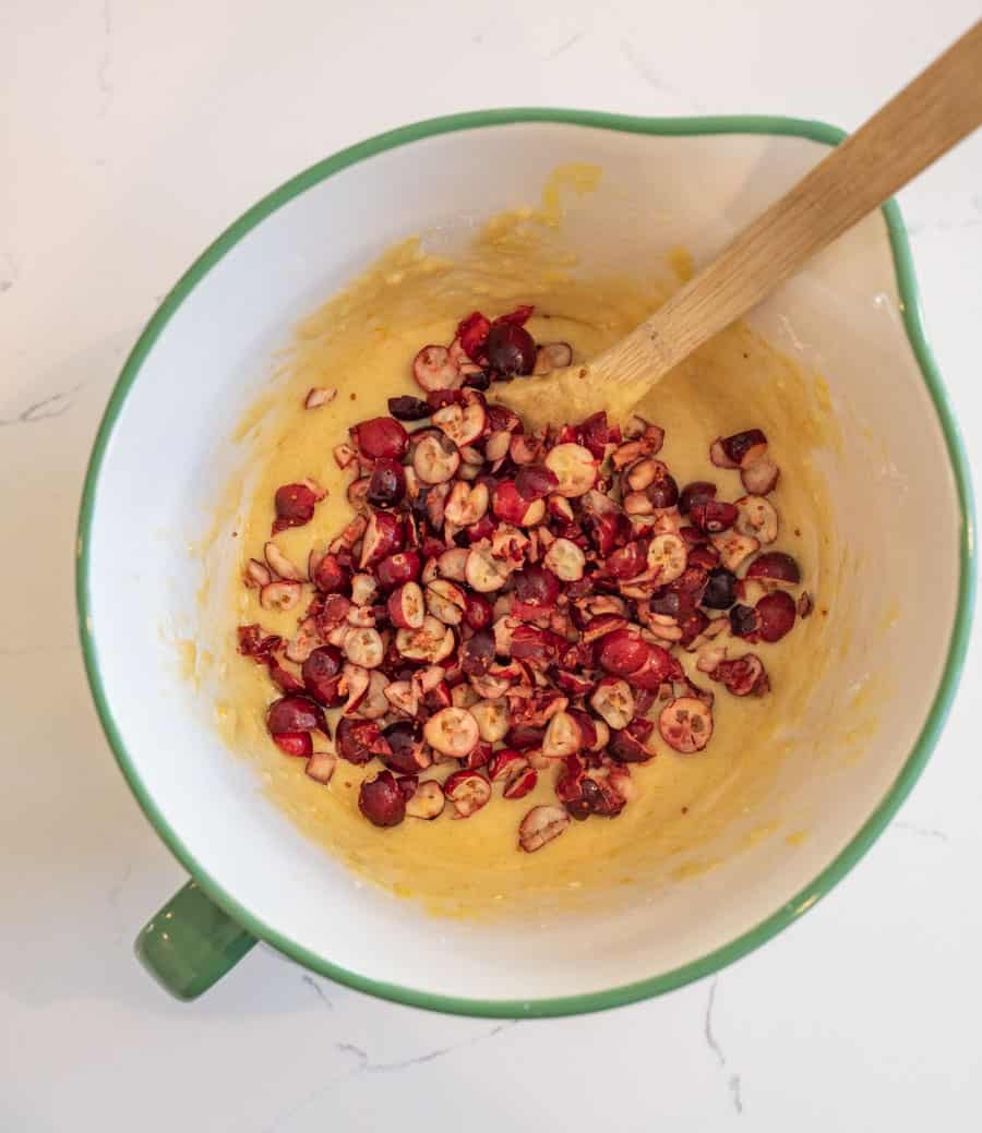 Chopped fresh cranberries being added to the batter for cranberry orange bread in a white mixing bowl with a green rim and a wooden spoon.