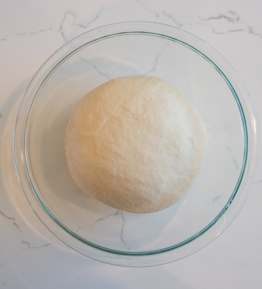 Ball of dough in a clear glass bowl on a white marble counter. 