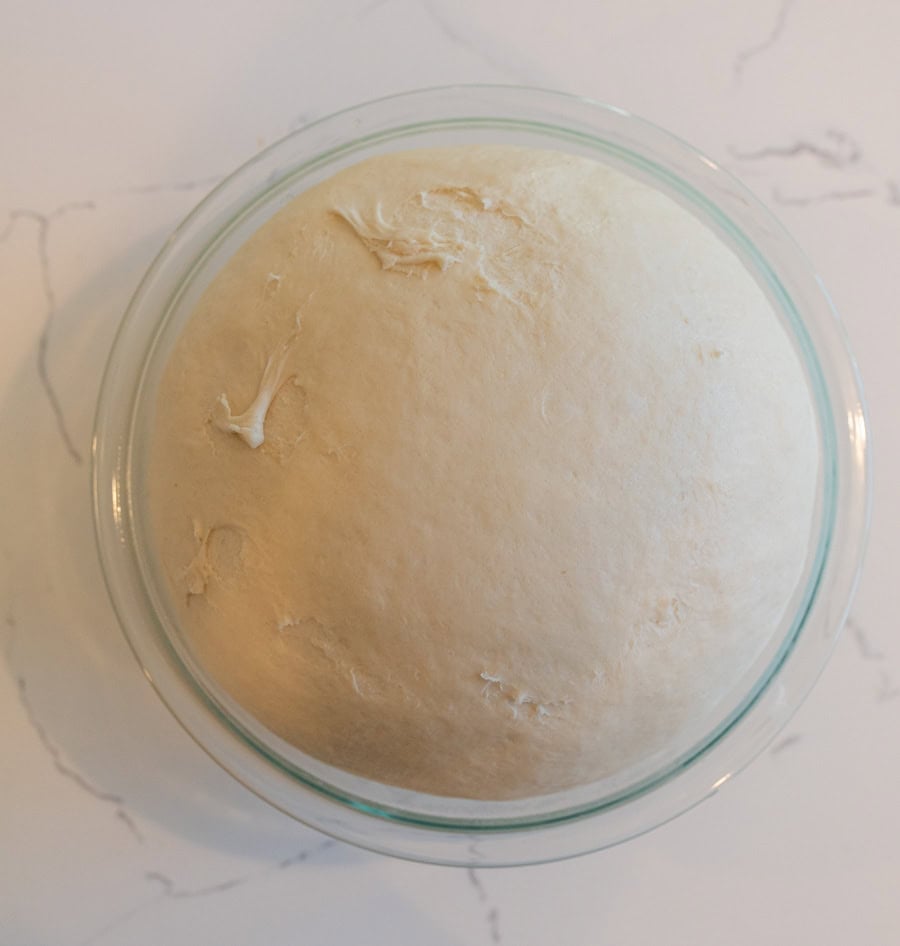 Ball of dough after rising in a clear glass bowl on a white counter.