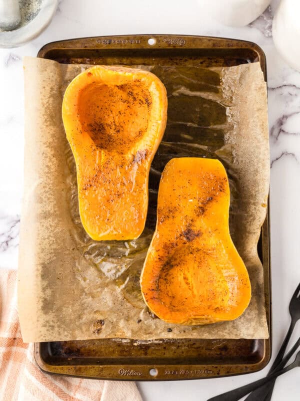 how to bake butternut squash showing the halved squash on parchment over a baking sheet seasoned and baked.