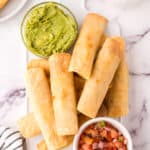 pile of taquitos on a plate with guacamole and salsa.