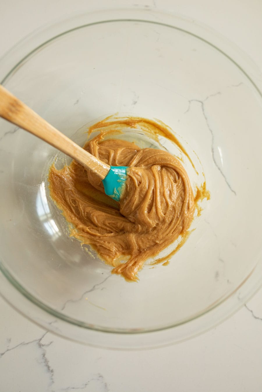 A clear glass mixing bowl with honey and peanut butter being mixed together with a spatula with a wooden handle. 