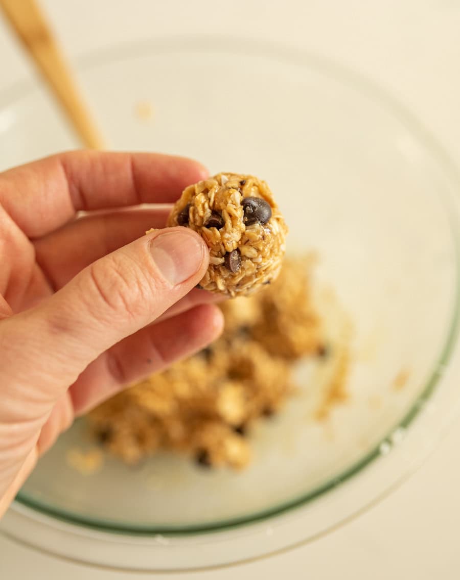 A hand holding a completed no-bake oatmeal energy ball with the clear glass bowl of dough in the background. 