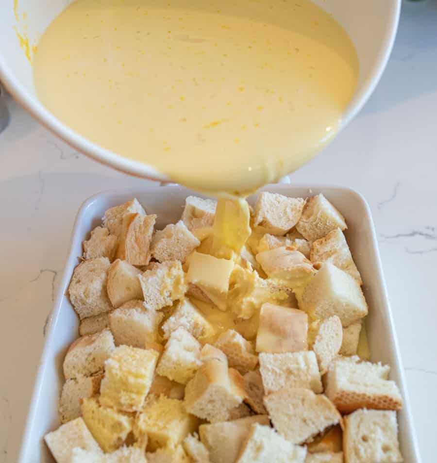 Egg and milk mixture being poured over the bread crumbs in a white baking dish. 