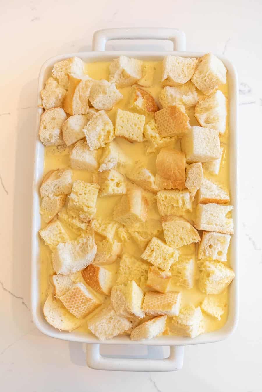 Bread cubes with wet ingredients in a white baking dish before being baked. 