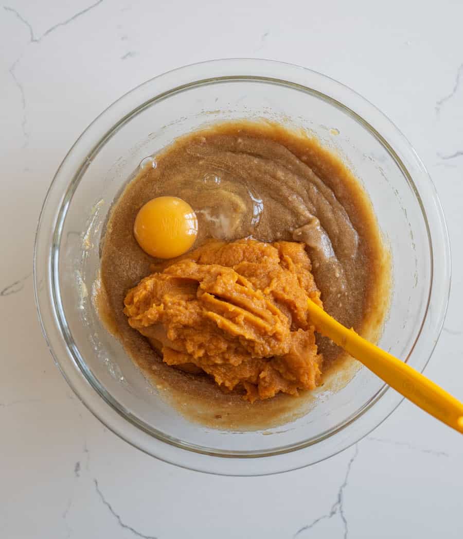 An egg and pumpkin added to the pumpkin cake batter in a clear glass bowl on a white counter. 