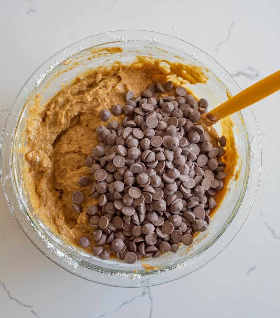 Chocolate chips added to the pumpkin cake batter in a clear glass bowl on a white counter. 