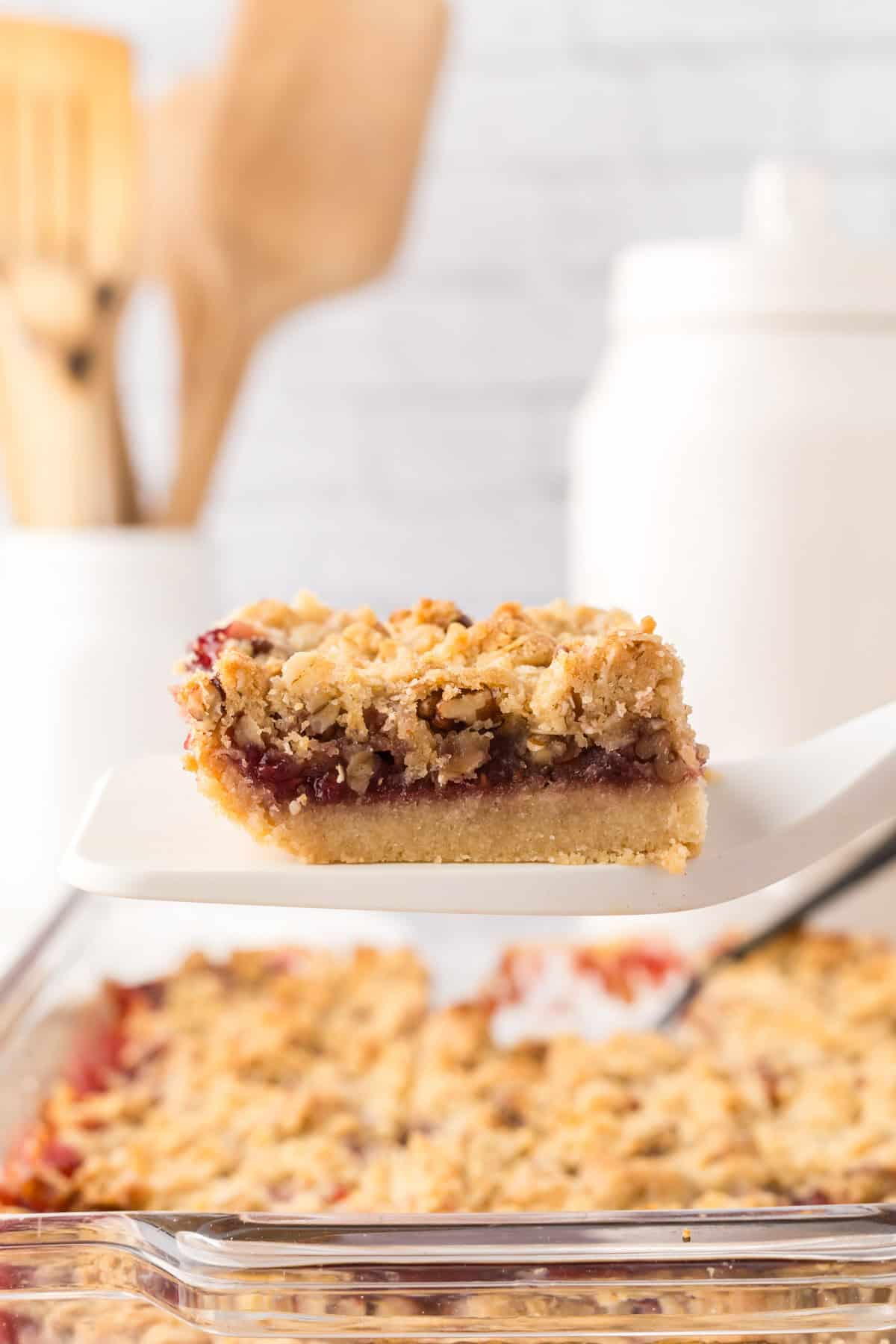 spatula with a serving held over clear glass dish with raspberry streusel bars.