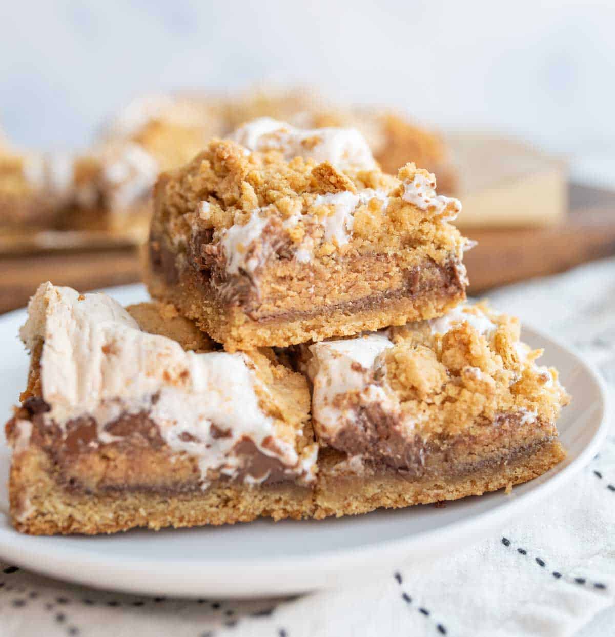 A plate with stacked dessert bars, featuring a crumbly golden crust, a layer of melted chocolate, and toasted marshmallows on top. The background shows a subtle checkered cloth and blurred wooden board with more dessert bars.