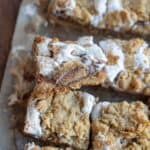 Close-up of Reeses Smores Bars on a tray, crowned with a crumbly layer and white icing. One piece is stacked on another, revealing a gooey, layered interior. The golden-brown bars boast visible chunks and a textured surface, capturing the essence of everyones favorite campfire treat.