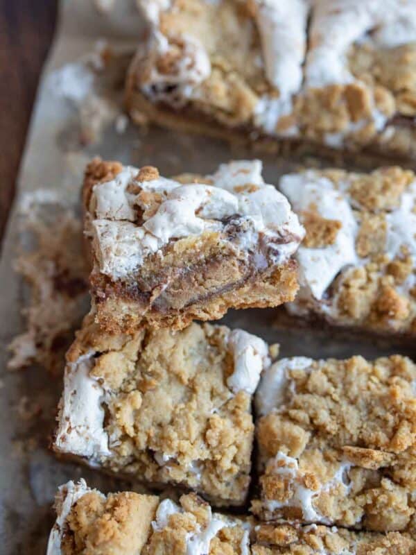 Close-up of Reeses Smores Bars on a tray, crowned with a crumbly layer and white icing. One piece is stacked on another, revealing a gooey, layered interior. The golden-brown bars boast visible chunks and a textured surface, capturing the essence of everyones favorite campfire treat.