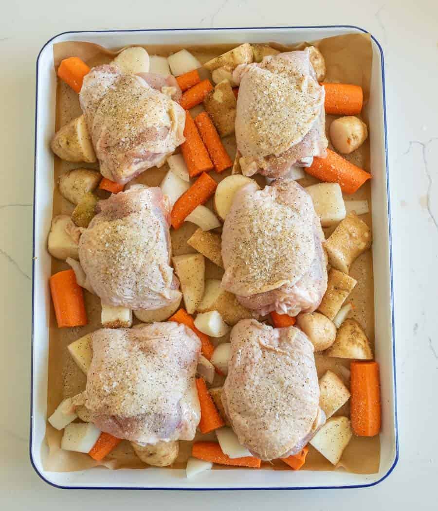 Raw chicken thighs on top of potatoes, onions, and carrots on a lined sheet pan ready to bake.