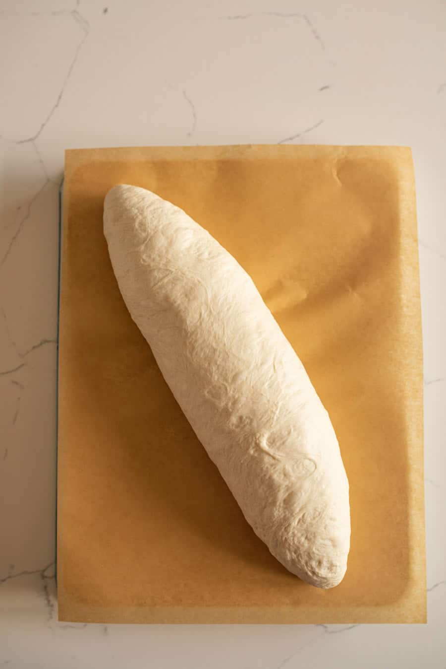 Italian bread dough on brown parchment paper after being shaped into a loaf. 