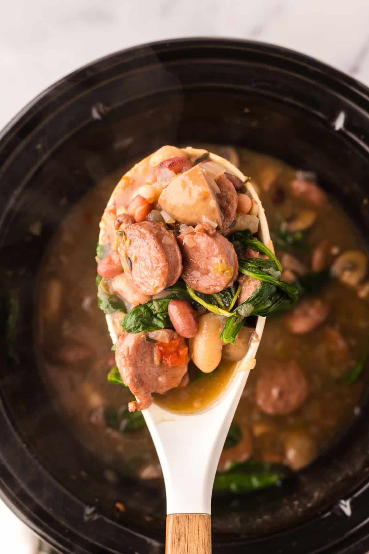 crockpot with sausage bean soup with spinach with being scooped with a white spoon.