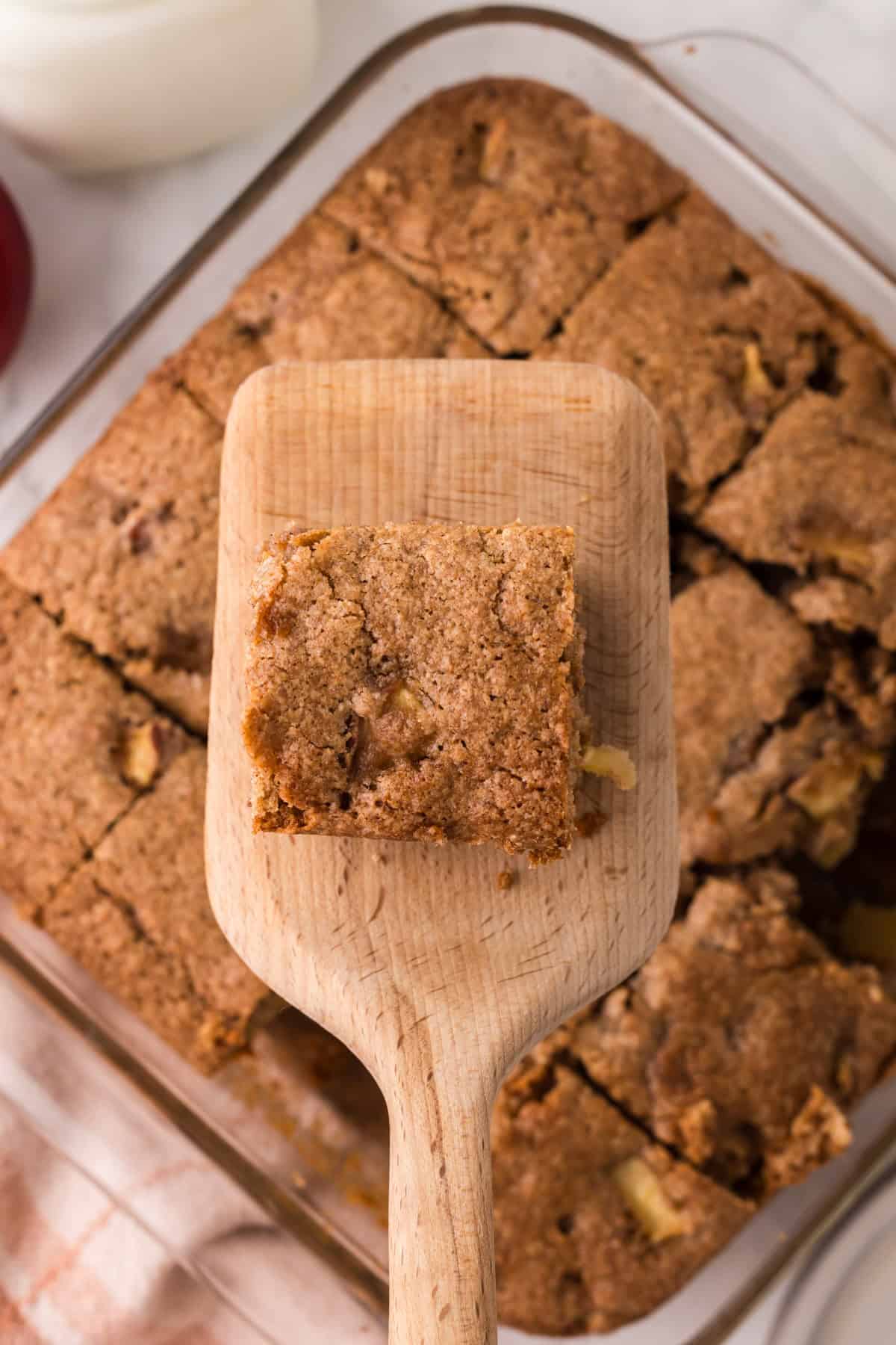 apple cake in a clear pan sliced into squares on a wooden spatula.