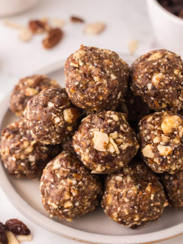 A plate stacked with several energy bites made of chopped nuts and dates, highlighting their coarse texture. Resembling the perfect recipe for cherry balls, the treats are round and set against a light background with scattered nuts visible around the plate.