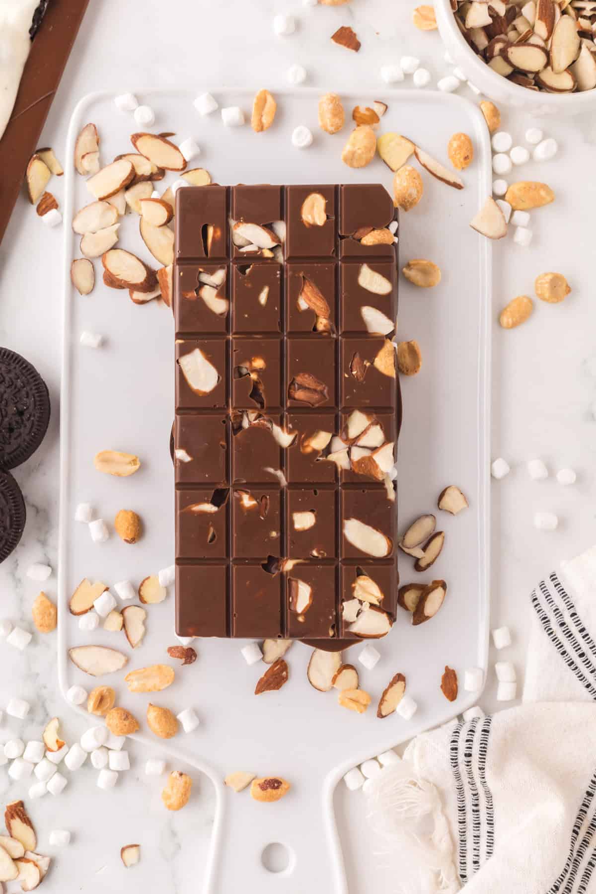 homemade candy bar with nuts and marshmallows surrounding it on a white cutting board.